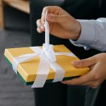 Nahaufnahme von Händen, die eine gelbe Geschenkbox mit einer weißen Schleife öffnen.