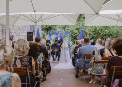 Trauungszeremonie im Freien mit Trauredner als Geistliche. Braut und Bräutigam sitzen, Gäste schauen zu.