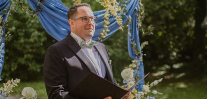 Freundlicher männlicher Hochzeitsoffiziant, Trauredner, hält das Zeremonienbuch am Hochzeitsbogen im Freien.