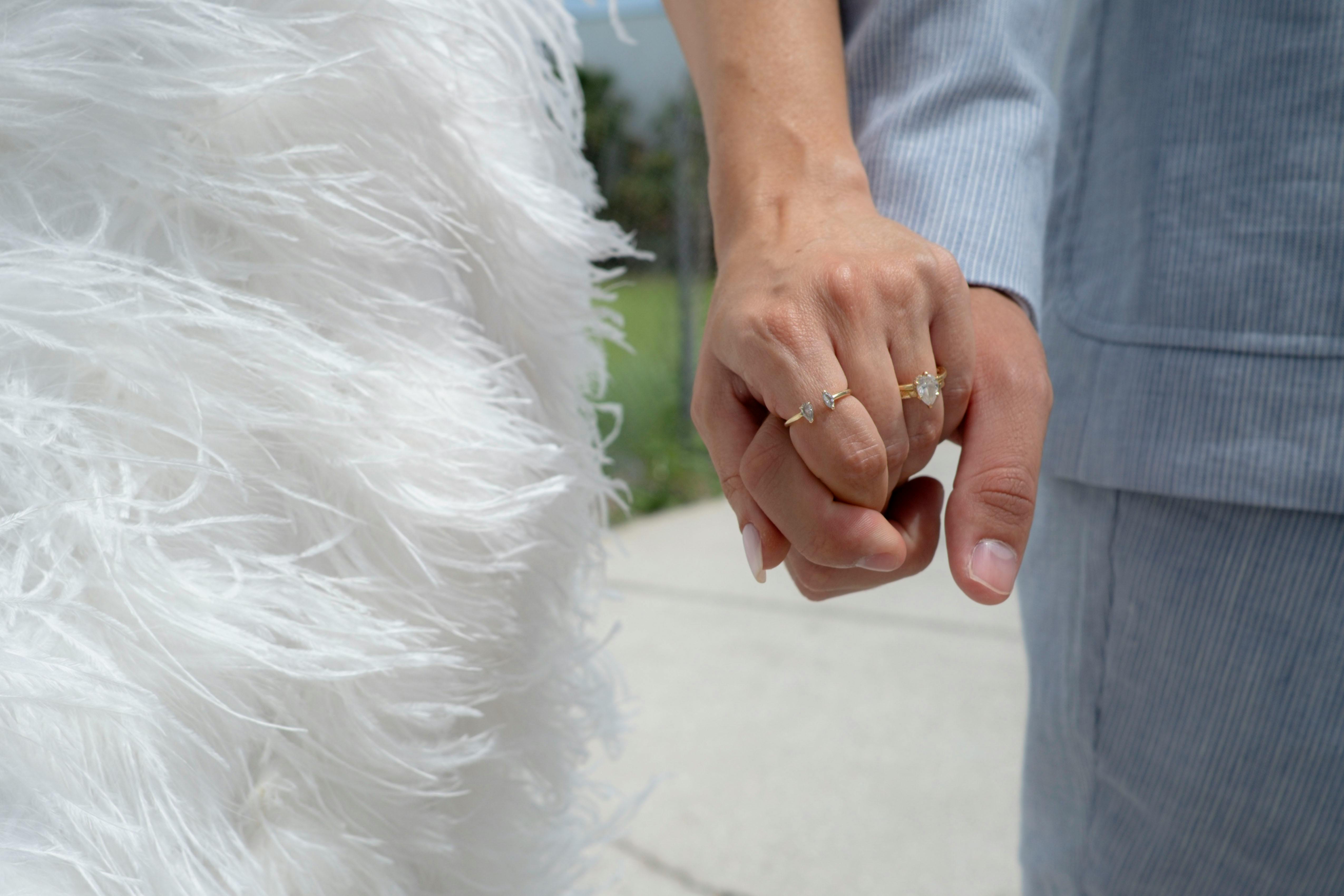 Brautpaar hält Hände mit Eheringen, Braut im weißen Kleid, Bräutigam im blauen Anzug.