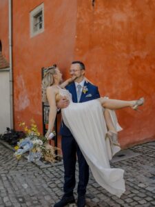 Bräutigam im blauen Anzug hält Braut im weißen Kleid vor orangefarbener Wand