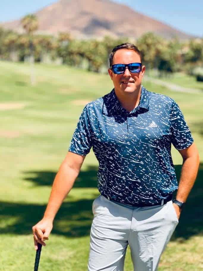 Mann mit Sonnenbrille hält einen Golfschläger auf einem sonnigen Platz