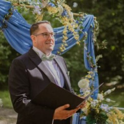 Freundlicher männlicher Hochzeitsoffiziant, Trauredner, hält das Zeremonienbuch am Hochzeitsbogen im Freien.