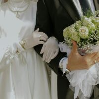 Braut und Bräutigam halten Händchen, sie trägt ein weißes Kleid und er hält einen Strauß weißer Rosen.